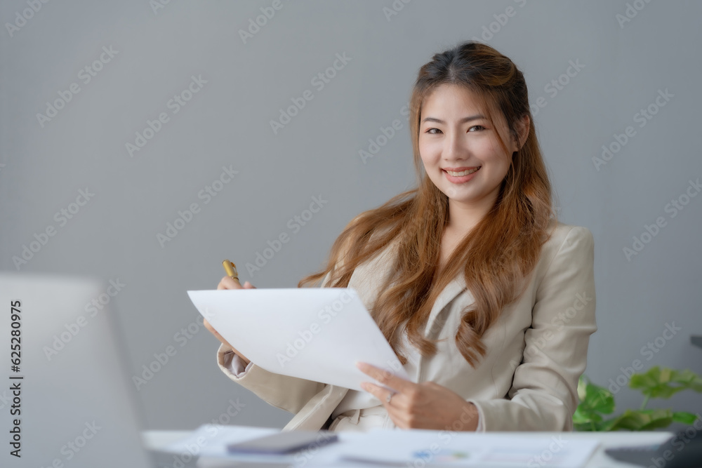 Happy Asian woman accountant at work sitting and taking note of the office report. Asian businesswoman writing a note making a report on investment profits earnings of a company. Happy Asian woman accountant at work sitting and taking note of the office report. Asian businesswoman writing a note making a report on investment profits earnings of a company.