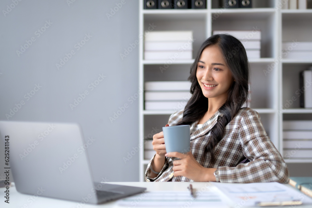 Successful Asian businesswoman smiling using laptop computer and holding coffee cup at office. Confident Asia businesswoman sitting happily in the office. Successful Asian businesswoman smiling using laptop computer and holding coffee cup at office. Confident Asia businesswoman sitting happily in the office.