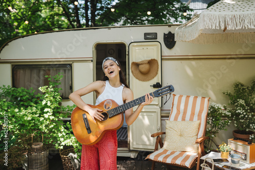 Happy hippie girl is having a good time with playing on guitar in camper trailer. Holiday, vacation, trip concept. photo