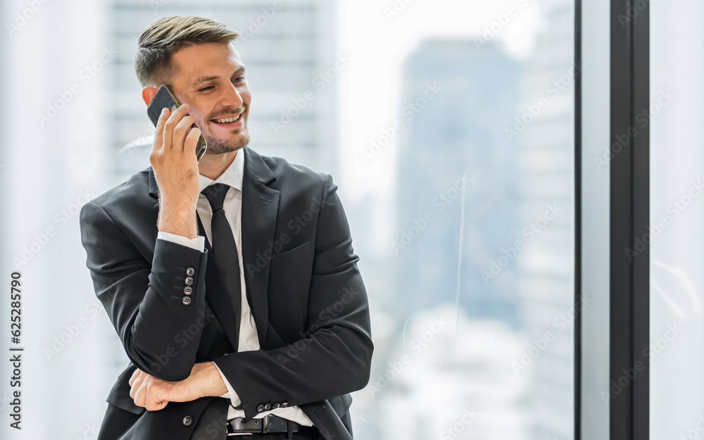 Handsome Caucasian business man wearing formal suit, necktie, smiling, standing beside window, using mobile phone, wifi for searching internet. Business, Communication, Technology, Networking Concept. Handsome Caucasian business man wearing formal suit, necktie, smiling, standing beside window, using mobile phone, wifi for searching internet. Business, Communication, Technology, Networking Concept.