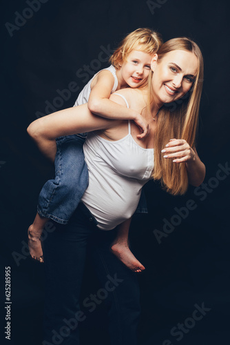 Pregnant girl with her daughter on her back, both smiling looking at the camera. Isolated on black.