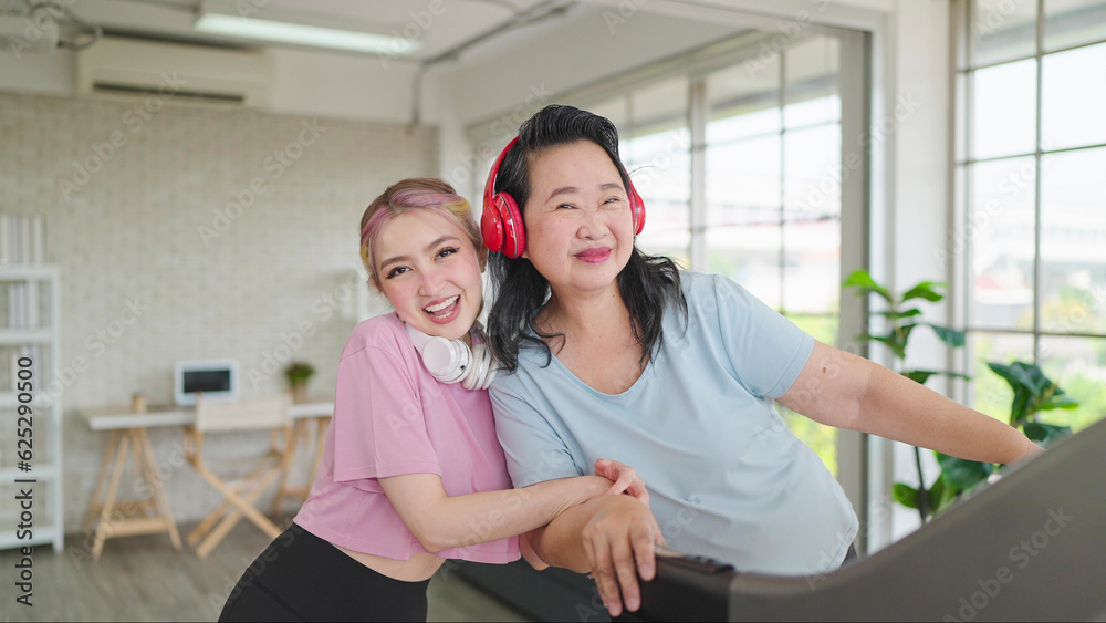 Happy asian elderly women and teenager daughter in sportswear wearing headphones while exercising at home. Smiling and looking at camera. Happy family, Health care concept Happy asian elderly women and teenager daughter in sportswear wearing headphones while exercising at home. Smiling and looking at camera. Happy family, Health care concept