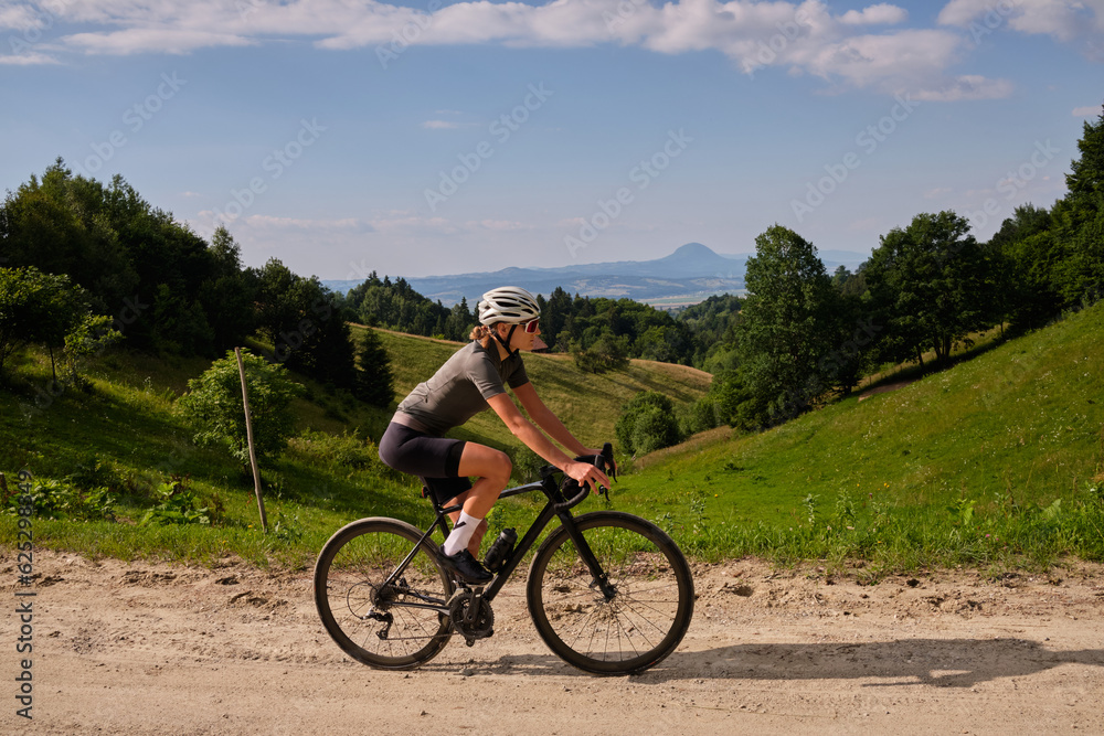 Female cyclist riding a gravel bike on a gravel road with a view of the mountains. Athlete is wearing cycling kit.