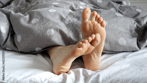 Closeup of a pair of feet belonging to a woman, visible from under the blankets on a cozy bed. Calm and comfortable lifestyle.