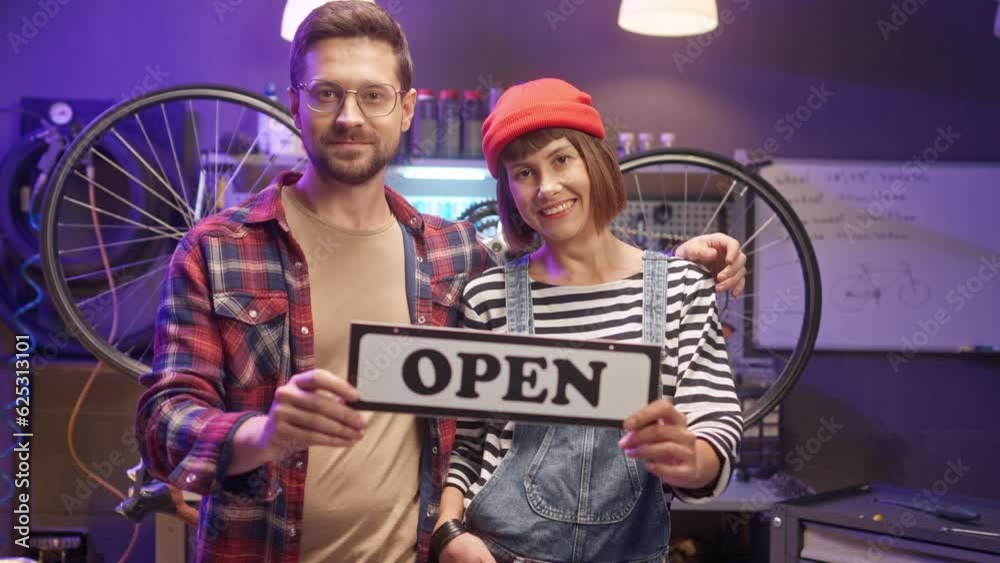Man and woman looking at camera and smiling. Two cheerful masters posing at workshop. Male and female holding sign with inscription open. Man and woman working at repair garage. Man and woman looking at camera and smiling. Two cheerful masters posing at workshop. Male and female holding sign with inscription open. Man and woman working at repair garage.