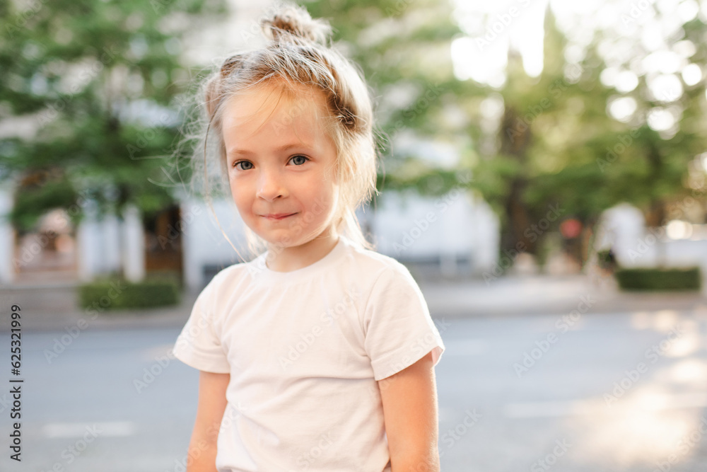 Cute kid girl 4-5 year old wear white t-shirt walking on city street outdoor. Looking at camera. Summer season. Childhood.