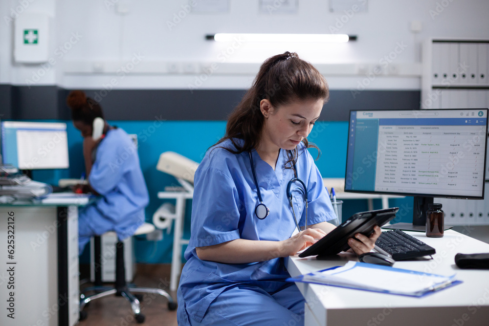 Young nurse checking appointments list on digital tablet in busy medical office. Adult woman healthcare specialist working with technology at hospital desk with stethoscope around neck - obrazy, fototapety, plakaty 