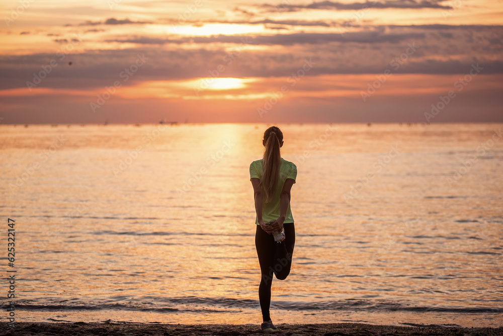 Young woman in sportswear is warming up early in morning on seashore at sunrise. Fitness
