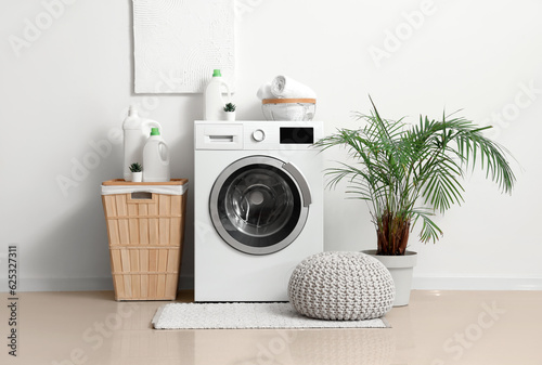Washing machine with bottles of detergent, laundry basket and pouf near white wall