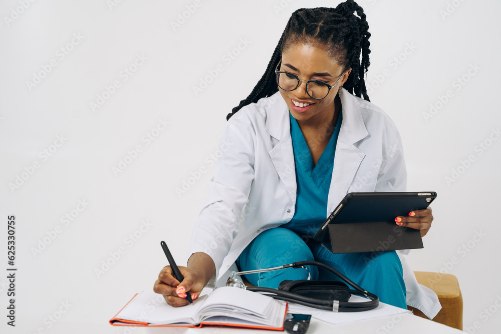 Smiling african american female doctor gp wears white medical coat using tablet at workplace gives remote online consultation, telemedicine chat Smiling african american female doctor gp wears white medical coat using tablet at workplace gives remote online consultation, telemedicine chat