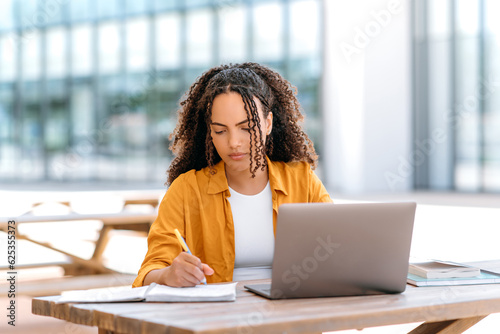 Focused trendy hispanic or brazilian curly haired girl, student or freelancer, sitting outdoors with a laptop, working or studying online, concentrated writing down information in her notepad photo