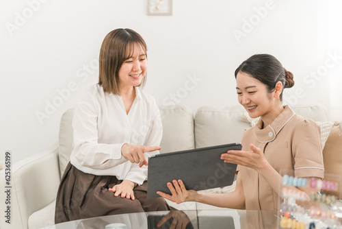 ネイルサロンでプランとデザインを説明するネイリストと見本を見ながらタブレットで選ぶ日本人女性
