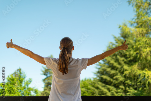 Young woman in casual clothing stands on porch with outstretched arms  enjoying clear sky.