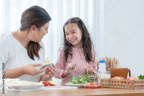Asian mother and daughter preparing breakfast in kitchen, cute child have fun mixing salad, mom cutting apples, milk, bread, tomatoes on table. Happy family and healthy food at home. Focus on kid