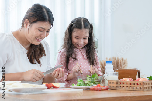 Asian mother and daughter preparing breakfast in kitchen, cute child have fun mixing salad, mom cutting apples, milk, bread, tomatoes on table. Happy family and healthy food at home. Focus on kid