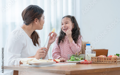 Asian mother and daughter preparing breakfast in kitchen, have fun mixing salad, mom peeling apples for cute child, milk, bread, tomatoes on table. Happy family and healthy food at home. Focus on kid