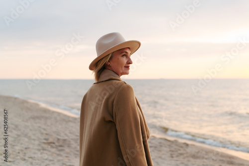 Obraz na plátně Lonely elegant senior woman in coat and hat standing on seashore at sunset in evening