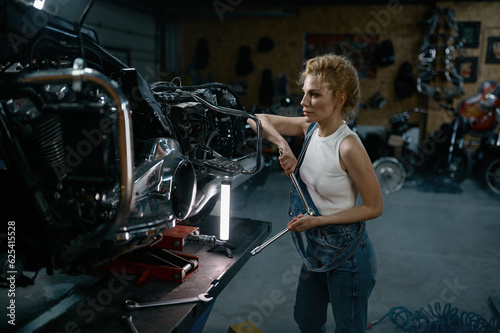 Talented girl wearing blue jumpsuit using work tool to repair motorcycle