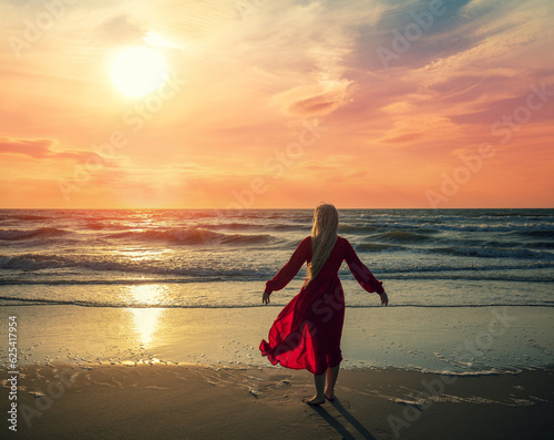 Seascape during orange sunrise with beautiful sky. Woman on the beach. Young happy woman in a long red fluttering dress walks on the seashore