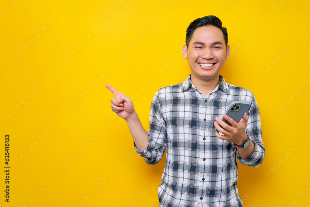Smiling happy handsome young Asian man wearing a white checkered shirt holding mobile phone and pointing a finger at empty space isolated over yellow background. People Lifestyle Concept Smiling happy handsome young Asian man wearing a white checkered shirt holding mobile phone and pointing a finger at empty space isolated over yellow background. People Lifestyle Concept