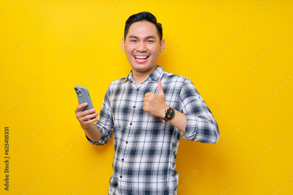 Smiling satisfied young Asian man wearing a white checkered shirt holding a mobile phone and showing thumbs up sign, recommends apps isolated over yellow background. People Lifestyle Concept Smiling satisfied young Asian man wearing a white checkered shirt holding a mobile phone and showing thumbs up sign, recommends apps isolated over yellow background. People Lifestyle Concept