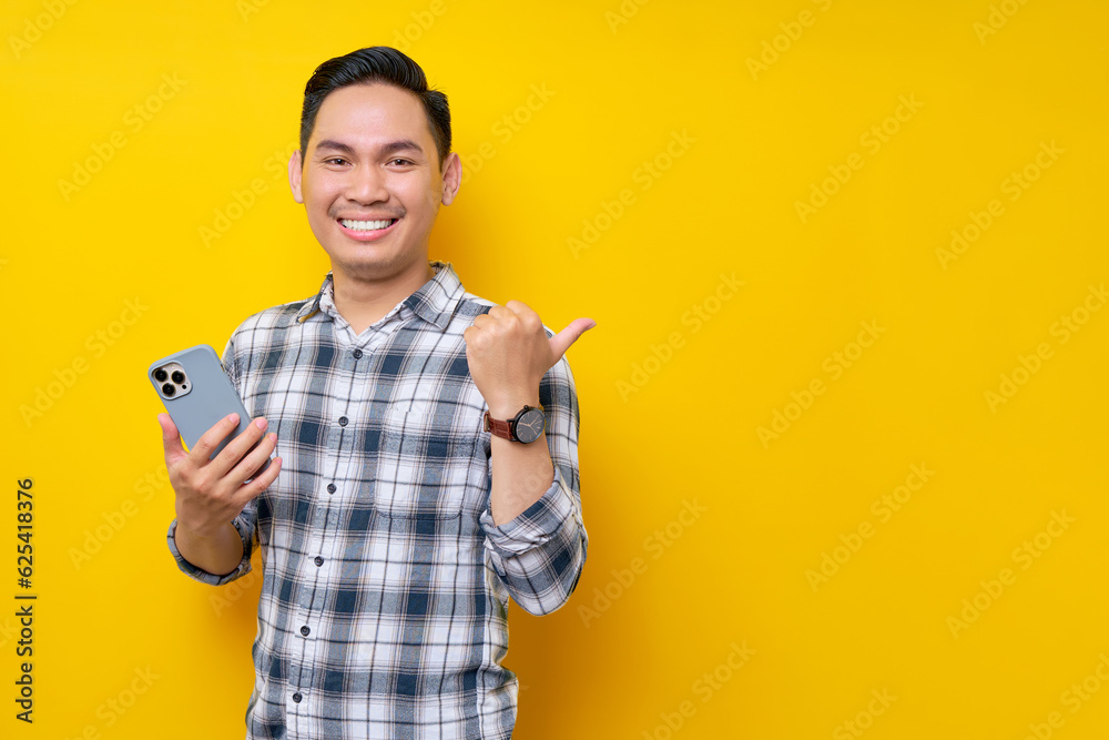 Smiling happy handsome young Asian man wearing a white checkered shirt holding a mobile phone and pointing thumb at empty space isolated over yellow background. People Lifestyle Concept Smiling happy handsome young Asian man wearing a white checkered shirt holding a mobile phone and pointing thumb at empty space isolated over yellow background. People Lifestyle Concept