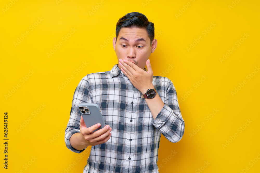 Shocked handsome young Asian man wearing a white checkered shirt cover mouth with hand and holding a smartphone isolated over yellow background. People Lifestyle Concept Shocked handsome young Asian man wearing a white checkered shirt cover mouth with hand and holding a smartphone isolated over yellow background. People Lifestyle Concept