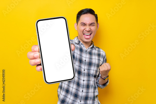 Smiling handsome young Asian man wearing a white checkered shirt showing a screen mobile phone for an advertisement and celebrating success isolated over yellow background. People Lifestyle Concept