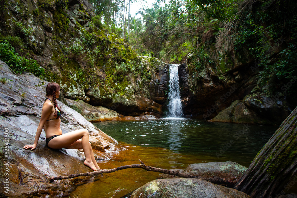 Fototapeta premium beautiful long hair woman in bikini sitting on the edge of a rock pool in front of large tropical waterfall at mount barney national park, queensland, australia 