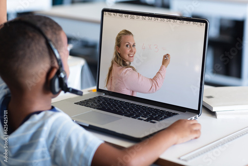 African american boy in headphones listening to female teacher teaching over video call on laptop