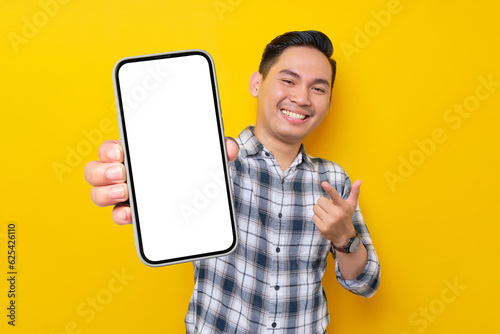 Smiling handsome young Asian man wearing a white checkered shirt pointing finger at mobile phone with blank screen, Advertising new mobile app isolated over yellow background. People Lifestyle Concept