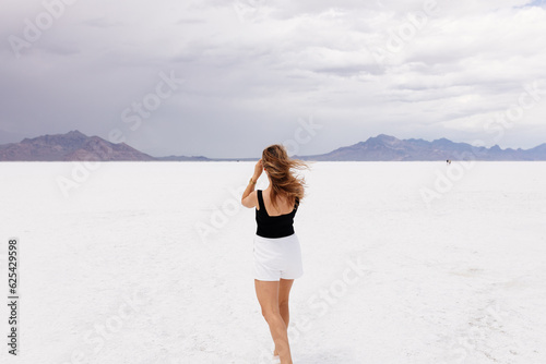 A beautiful unusual landscape on which a large desert of white salt and blue mountains in the distance on a sunny clear day. A young woman in white shorts and black top walks in desert of white salt. 