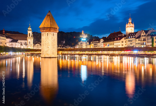 Night time panorama of Luzern old town in Switzerland. Colorfully illuminated buidings reflected in river Reuss (Lake Lucerne) in blue hour twilight. Famous landmark wooden ”Chapel bridge“ with tower.
