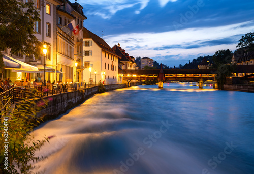 Night time panorama in Lucerne old town in Switzerland. Colorfully illuminated waterfront of river Reuss with strong current in evening blue hour twilight. Wooden bridge, cafes and city wall tower.