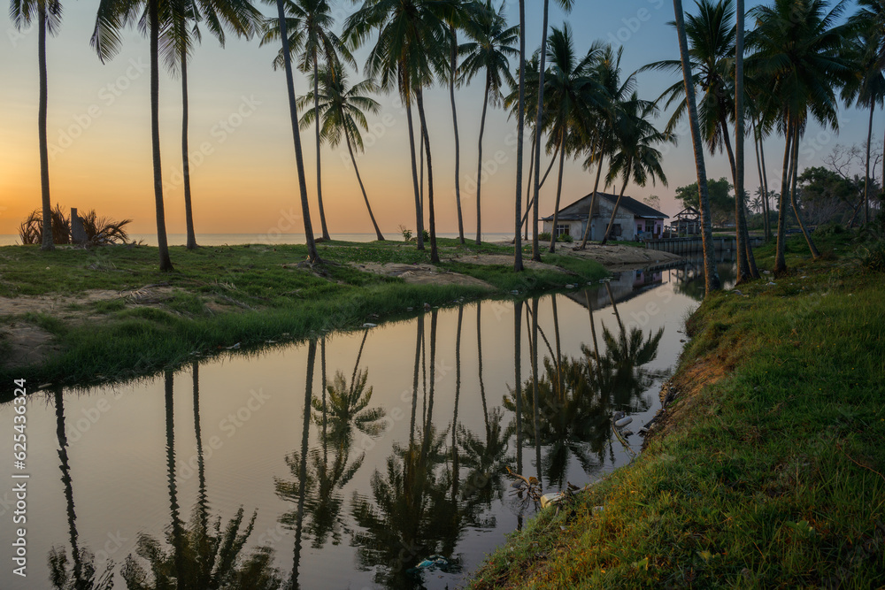 Sunrise over the beautiful beach with full of coconut trees and a small hut with reflection 