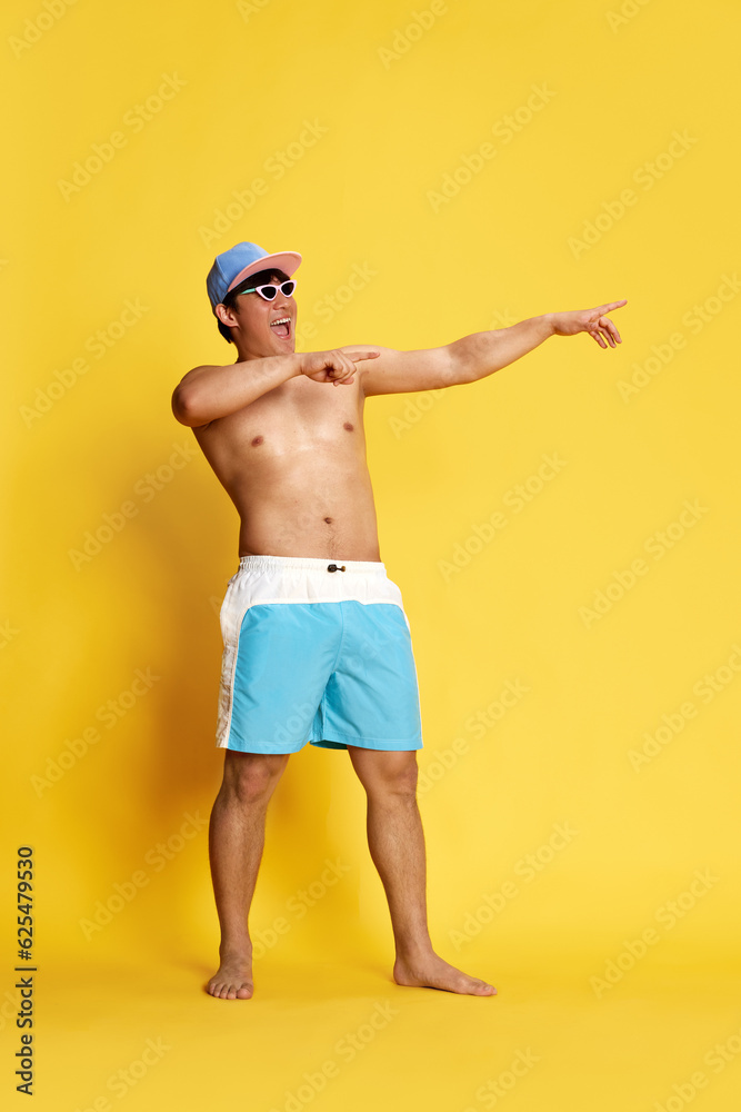Full-length portrait of joyful, cheerful young man in swimming trunks, sunglasses and cap posing over yellow studio background. Concept of summer, vacation, leisure time, holidays, human emotions, ad