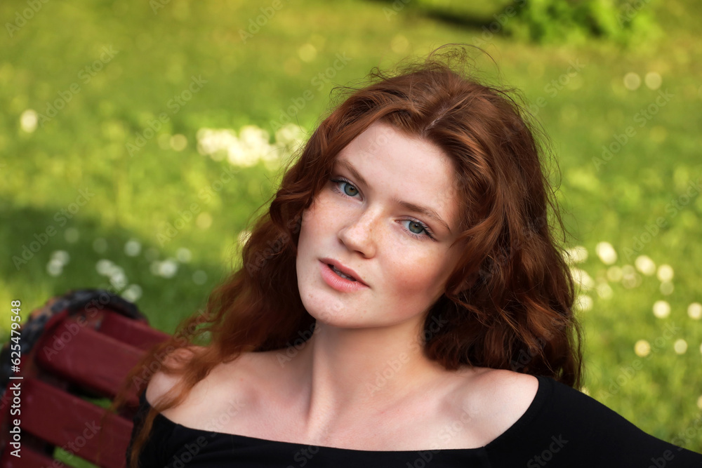 Attractive girl with long red hair sitting on a bench in summer park. Young sensual model, female beauty Attractive girl with long red hair sitting on a bench in summer park. Young sensual model, female beauty