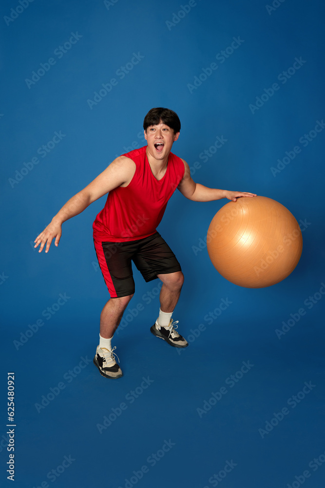 Full-length portrait of young ma in sportswear training, playing with fitness ball against blue studio background. Concept of sport, fitness, active and healthy lifestyle, human emotions, ad Full-length portrait of young ma in sportswear training, playing with fitness ball against blue studio background. Concept of sport, fitness, active and healthy lifestyle, human emotions, ad