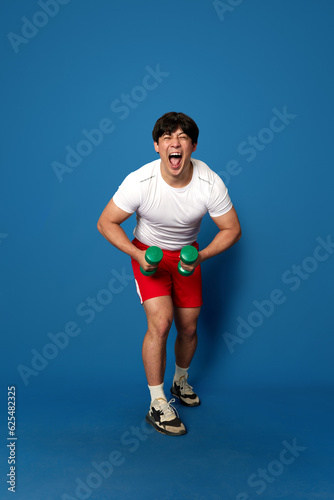 Full-length portrait of young man in sportswear training with dumbbells and having fun against blue studio background. Concept of sport, fitness, active and healthy lifestyle, human emotions, ad