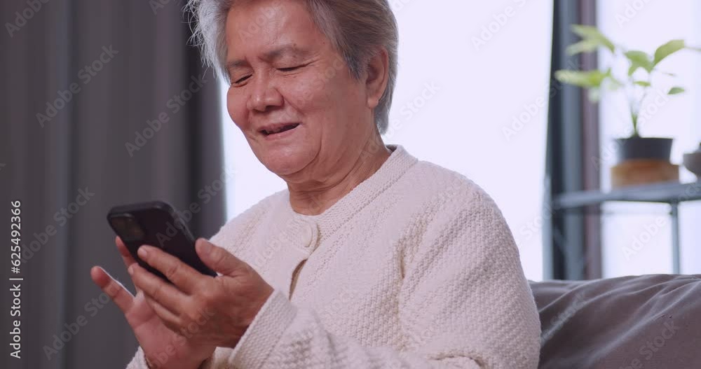 Happy Asian old woman using smart phone while sitting on sofa at home.