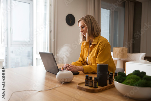 woman working on laptop from home near essential oil aroma diffuser humidifier diffusing water articles in the air. Aroma therapy photo