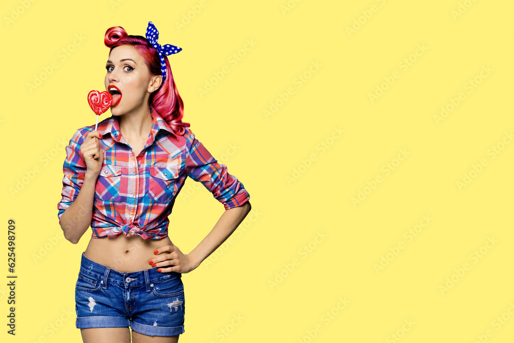 Portrait of beautiful woman licking heart shape lollipop dressed in pinup style plaid shirt, isolated on yellow  background. Purple red girl in retro fashion and vintage studio concept. Mock up.