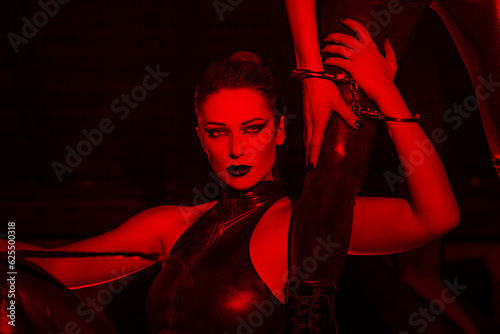 Sexy dominatrix in latex catsuit holding lovers leg in red light