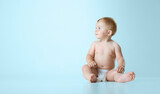 Beautiful little baby, toddler in diaper sitting calmly on floor and looking away against light blue studio background. Concept of childhood, newborn lifestyle, happiness, care. Copy space for ad