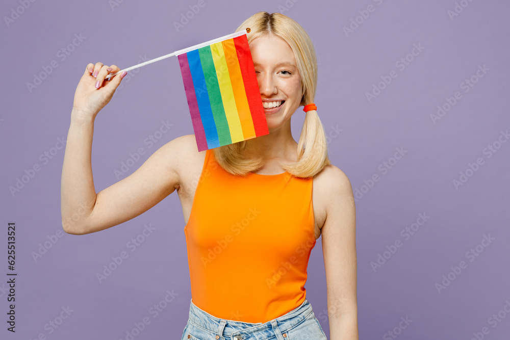 Young blonde lesbian woman she wear orange tank shirt casual clothes hold in hand cover eye with striped flag isolated on plain pastel light purple background. Pride day june month love LGBTQ concept. Young blonde lesbian woman she wear orange tank shirt casual clothes hold in hand cover eye with striped flag isolated on plain pastel light purple background. Pride day june month love LGBTQ concept.