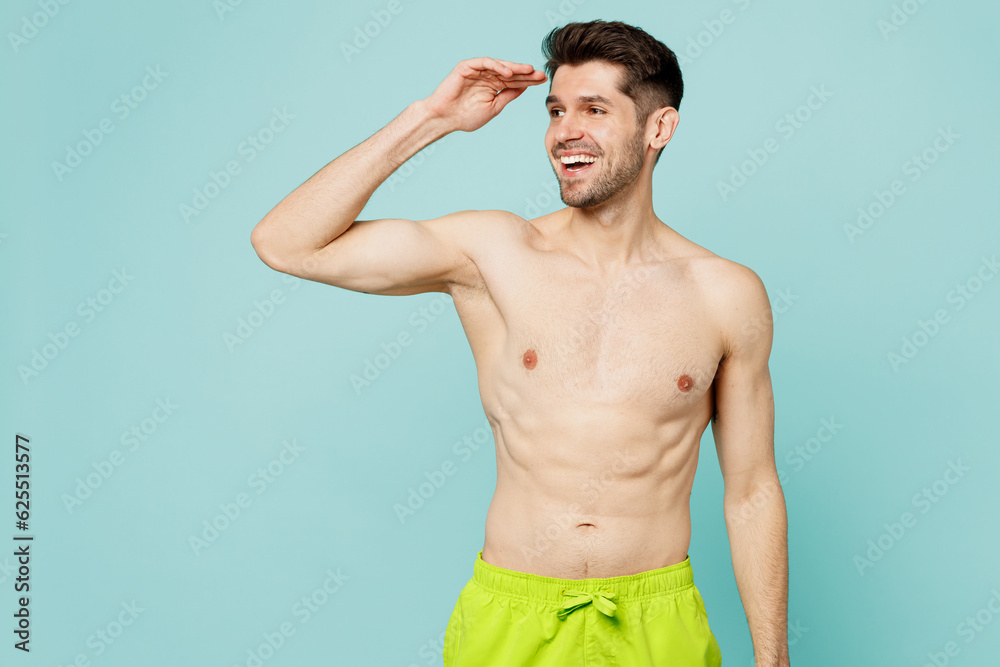 Young fun sexy man wear green shorts swimsuit relax near hotel pool hold hand at forehead look far away distance isolated on plain light blue cyan background. Summer vacation sea rest sun tan concept.