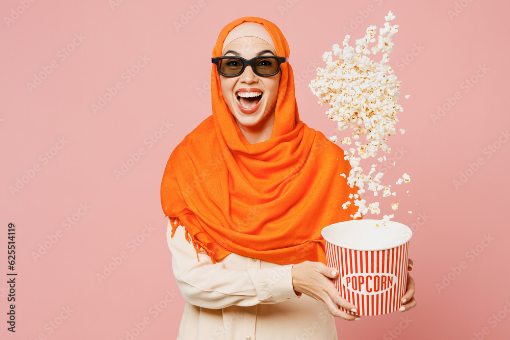Obraz premium Young arabian muslim woman in 3d glasses wear orange abaya hijab watch movie film hold bucket toss up popcorn in cinema isolated on plain pink background. Uae middle eastern islam religious concept.