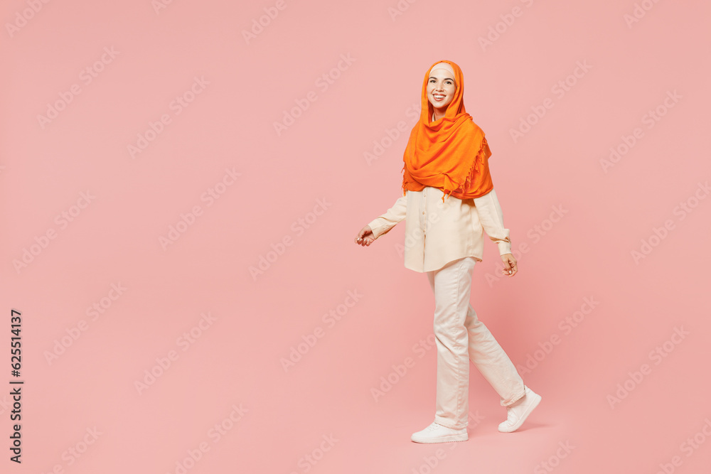 Full body side profile view smiling young arabian asian muslim woman wear orange abaya hijab walk go strolling look camera isolated on plain pink background Uae middle eastern islam religious concept Full body side profile view smiling young arabian asian muslim woman wear orange abaya hijab walk go strolling look camera isolated on plain pink background Uae middle eastern islam religious concept
