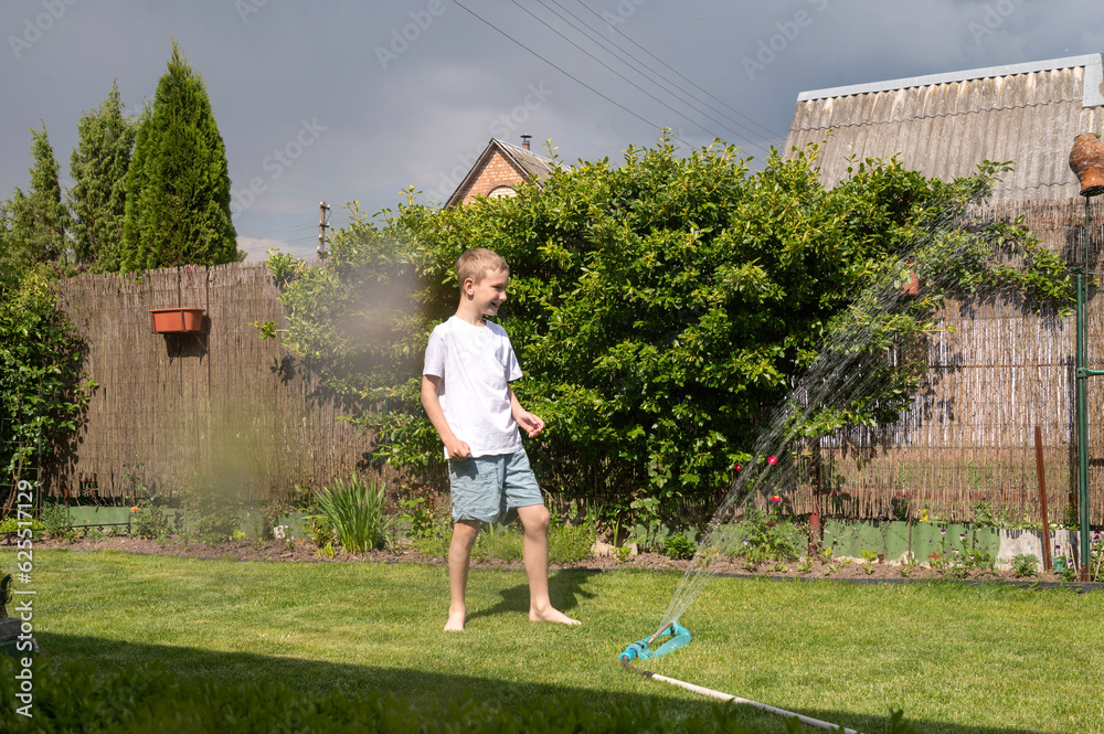 A cute boy is standing on the lawn under drops of water. Running under the sprinkler