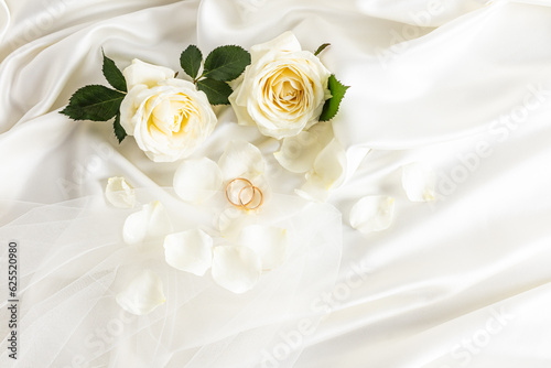 Two gold wedding rings on a beige satin background with live tea roses and part of the bride's veil. Chic layout for design. Wedding background